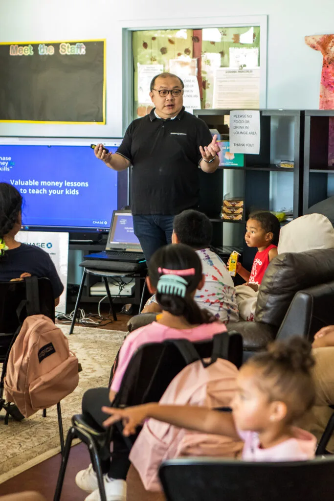A man gestures while standing next to a TV screen that reads "Valuable money lessons to teach your kids."
