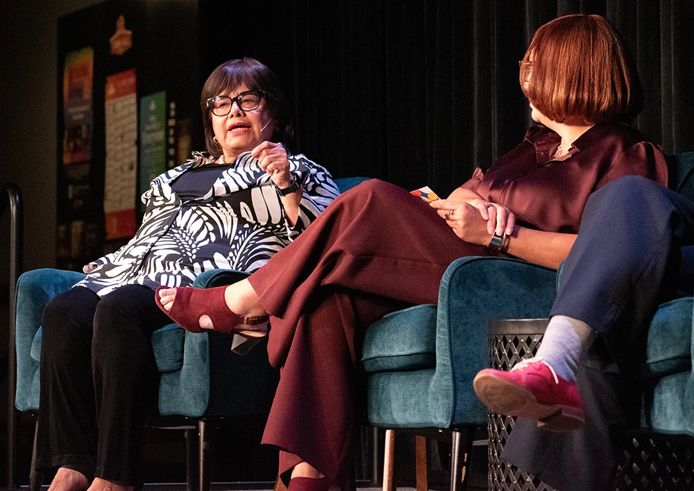 A woman seated in a chari gestures as she speaks while another woman looks on.