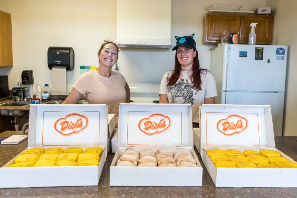 Two smiling women stand behind three boxes filled with hamburgers labeled "Dick's."