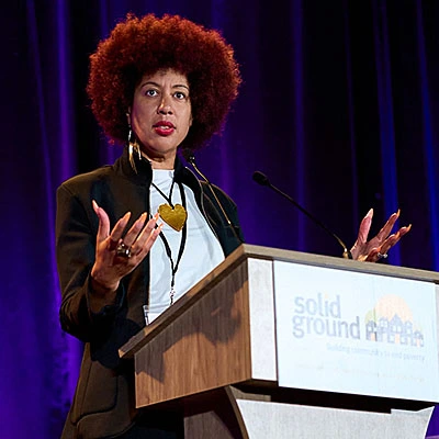 Woman with an afro wearing white shirt and dark jacket gestures as she speaks at a podium on a stage.