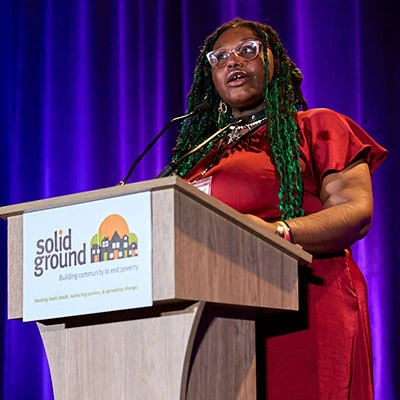 A woman in a red dress and long teal dreadlocks speaks at a podium on a stage.