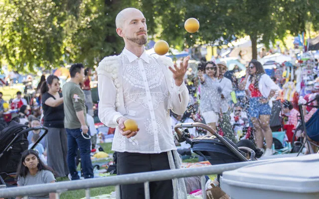 A man juggles three balls at Downtown Park while many people enjoy themselves behind him.