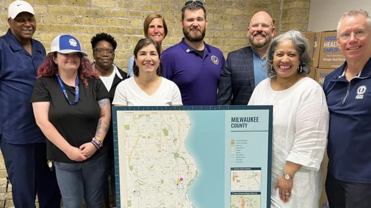 A diverse group of eight people stand smiling indoors behind a large Milwaukee County map