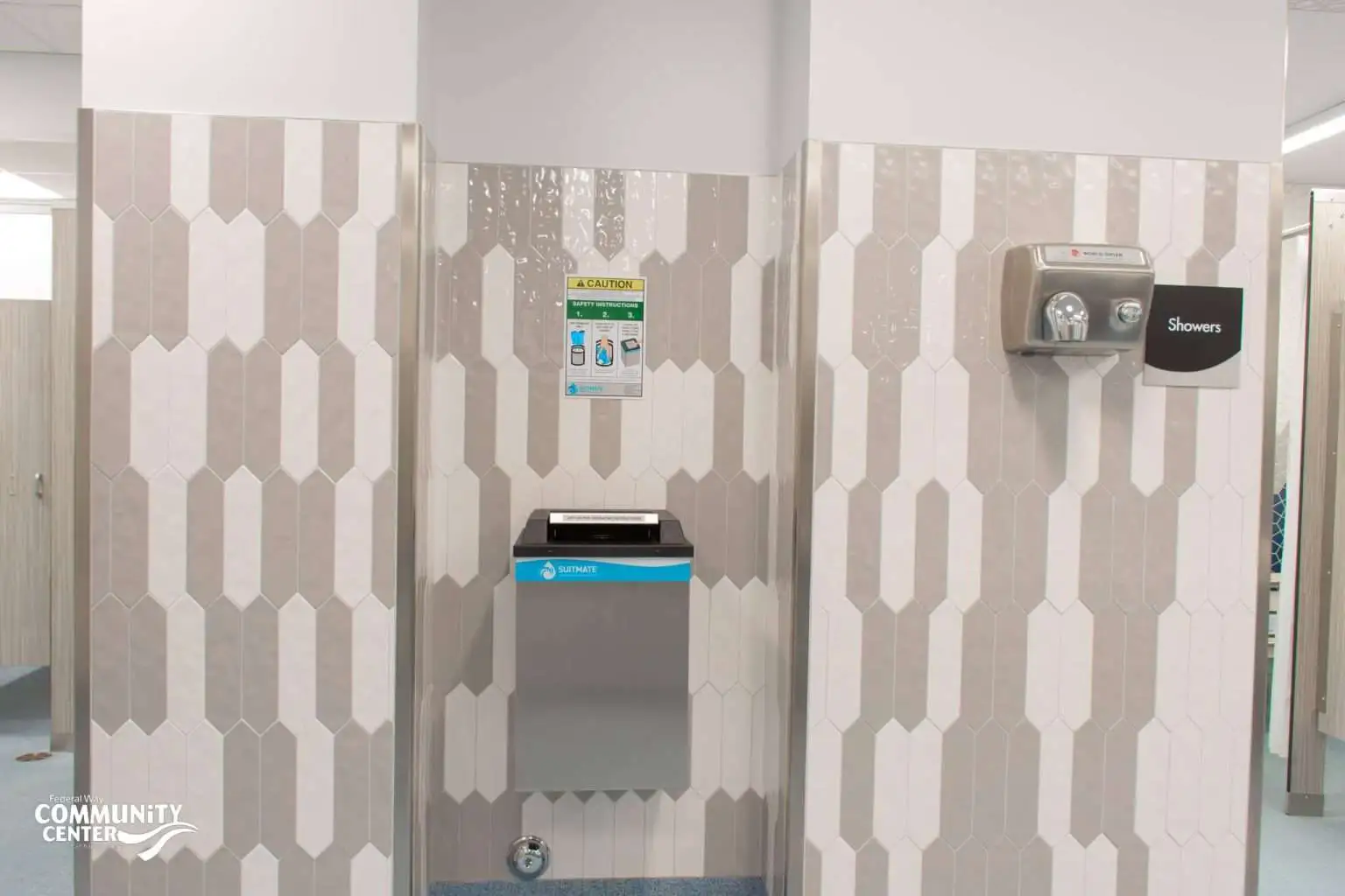 FWCC locker room area with beige and white vertical tile, a wall-mounted suit spinner machine, a caution sign, and a hand dryer near the entrance to the showers.