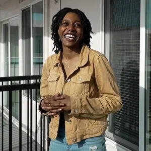A young woman in a tan jacket poses on her apartment deck, with a beaming smile on her face and her hands clasped.