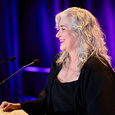 A woman with long silver hair stands at a microphone.