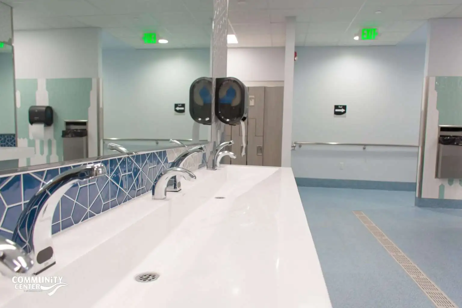 Renovated FWCC locker room with a long white sink counter, chrome faucets, blue geometric tile backsplash, light blue flooring, lockers in the background, and wall-mounted paper towel dispensers.