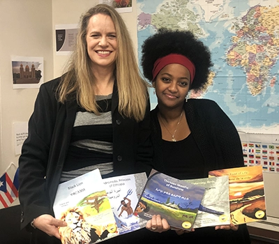 A woman and her teenage daughter hold a selection of colorful picture books infront of a map of the world.