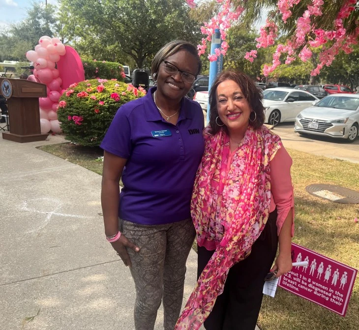 Two women smiling outdoors at a breast cancer awareness event. One is wearing a purple shirt, the other a pink scarf. Pink flowers and balloons adorn the background.