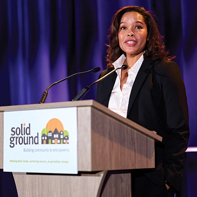 A woman in a dark suit jacket stands at a podium with a sign that reads 