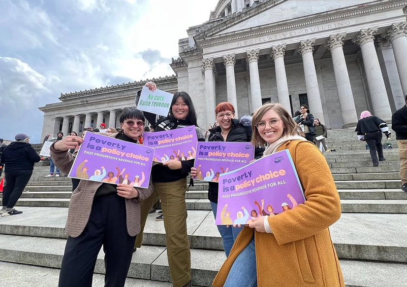 Four people standing on steps infront of a state legislative building holding signs that read 