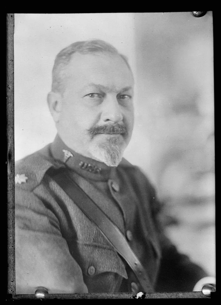 A man with a mustache and goatee wears a military uniform with shoulder insignia, looking confidently at the camera.
