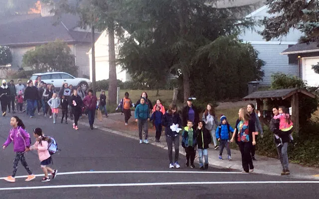 A large group of schoolchildren and parents walk in a procession down a neighborhood street and on a crosswalk.