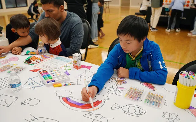A boy draws a rainbow. At the table next to him, a man draws with two younger children leaning on him and watching him.