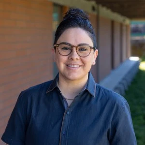 This is a headshot or Dalila. She is wearing a dark blue shirt and brown tortoiseshell glasses.