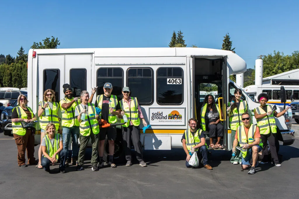 A group of people wearing yellow safety vests and carrying various cleaning supplies crowd around a bus labeled "Solid Ground."