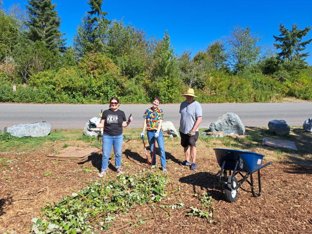 Three volunteers pose next to a wheelbarrow and a fresh layer of mulch.