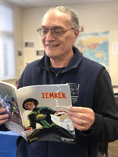 A man with glasses and a blue jacket smiles as he reads from a comic book titled "Jember."