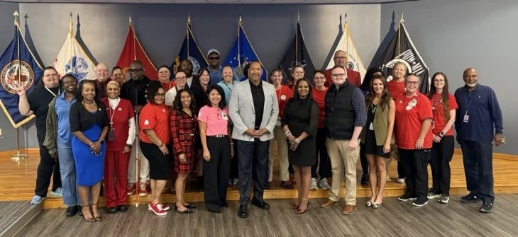 A diverse group of 27 people stand smiling on a wooden stage. Behind them are military flags displayed on the wall.