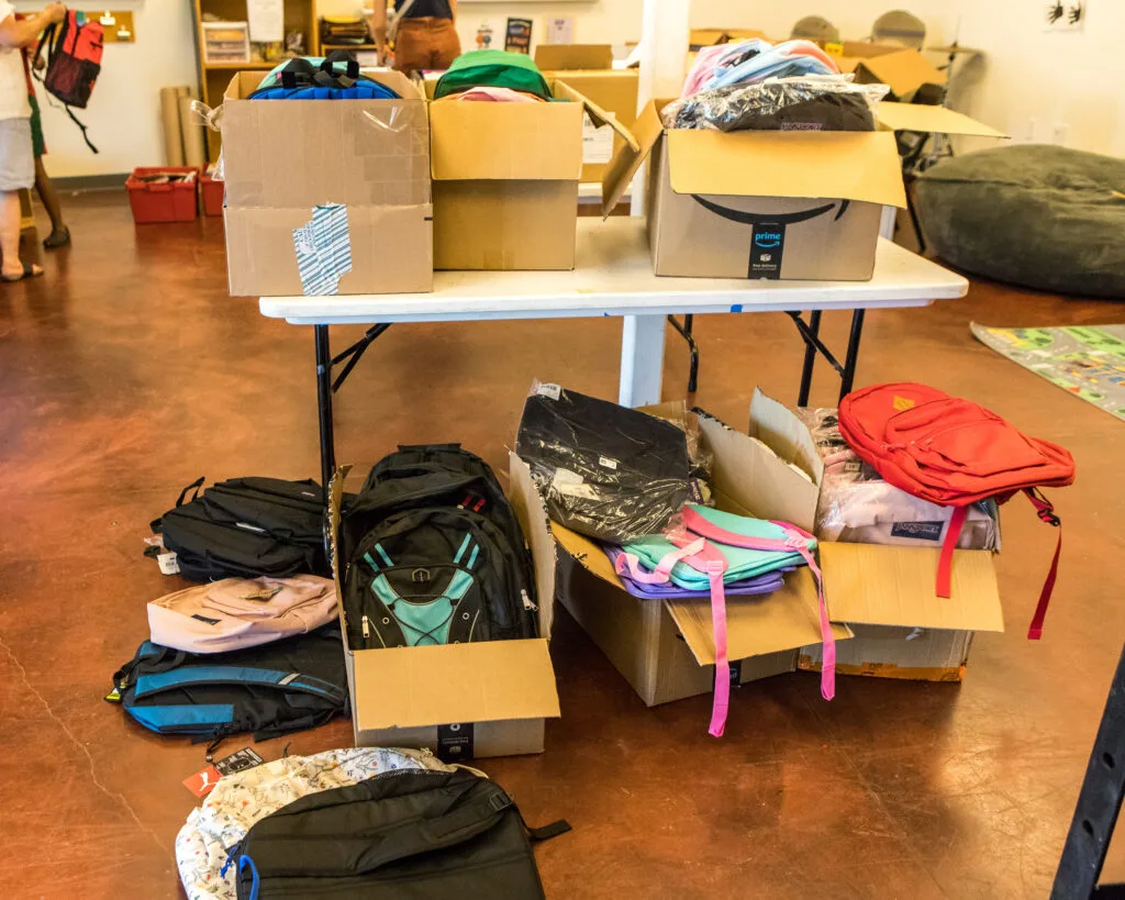 Cardboard boxes filled with backpacks sit on and under a folding table.