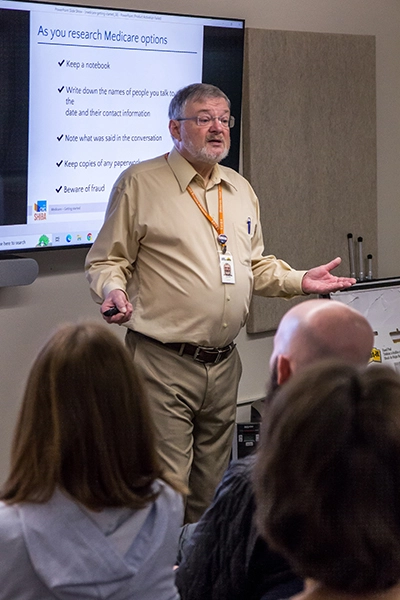 A man giving a presentation stands in front of a projection that reads 