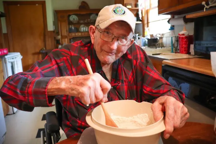Elderly man in a plaid shirt and cap stirs flour in a bowl at a cozy kitchen. He smiles in a homey and inviting scene.