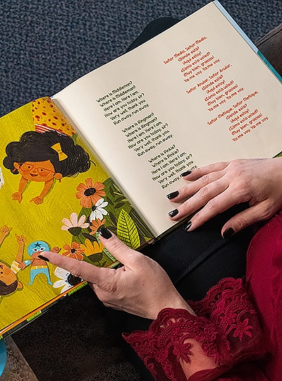 Hands of a woman holding an open picture book. She wears black nail polish and a red blouse.