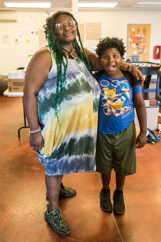 A woman with green braids and wearing a tie-dyed dress puts her arm around her young son, who has a mock shocked expression on his face.