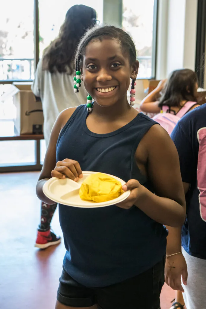 A girl holding a plate of food smiles into the camera.