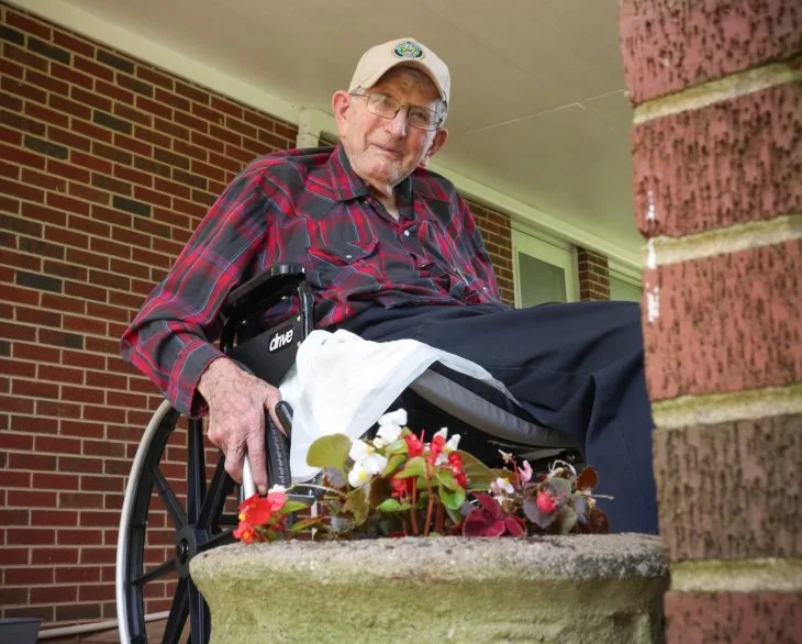 Elderly man in a plaid shirt and cap, sitting in a wheelchair on a porch. He smiles softly next to a pot of colorful flowers, brick wall in the background.