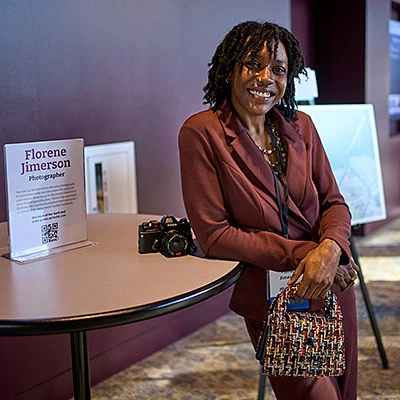 A young woman in a brown suit smiles and leans against a round table in a convention center lobby.