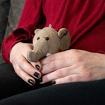 Hands of a woman holding a small stuffed elephant. She wears black nail polish, black pants, and a red blouse.