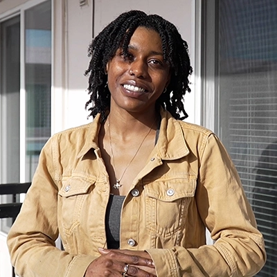 Portrait of a young woman in a tan jacket on her apartment deck, with a beaming smile on her face and her hands clasped.