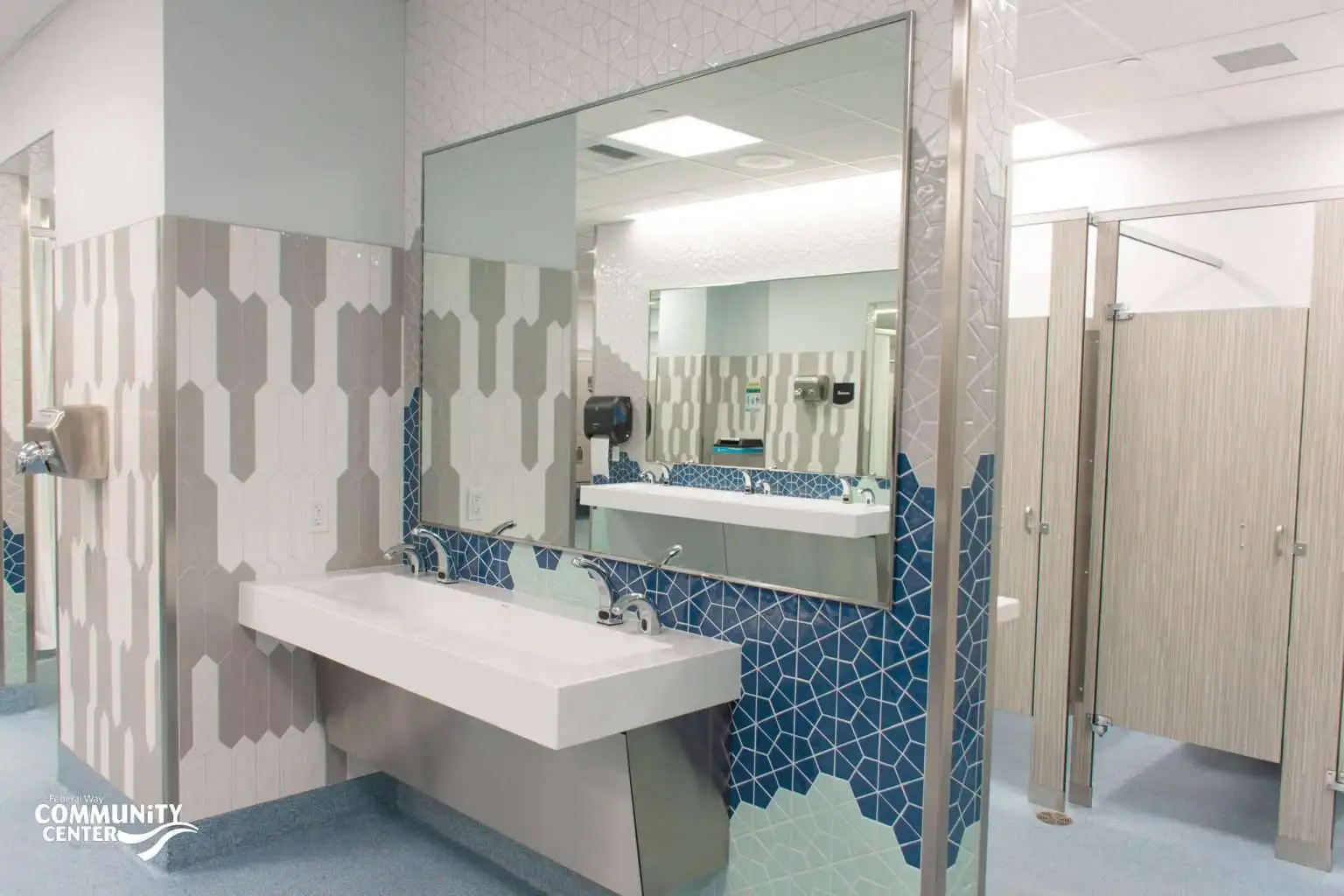 FWCC locker room sink area with a large mirror, white sink, blue geometric tile backsplash, and beige and white vertical tile on surrounding walls; restroom stalls visible in the background.