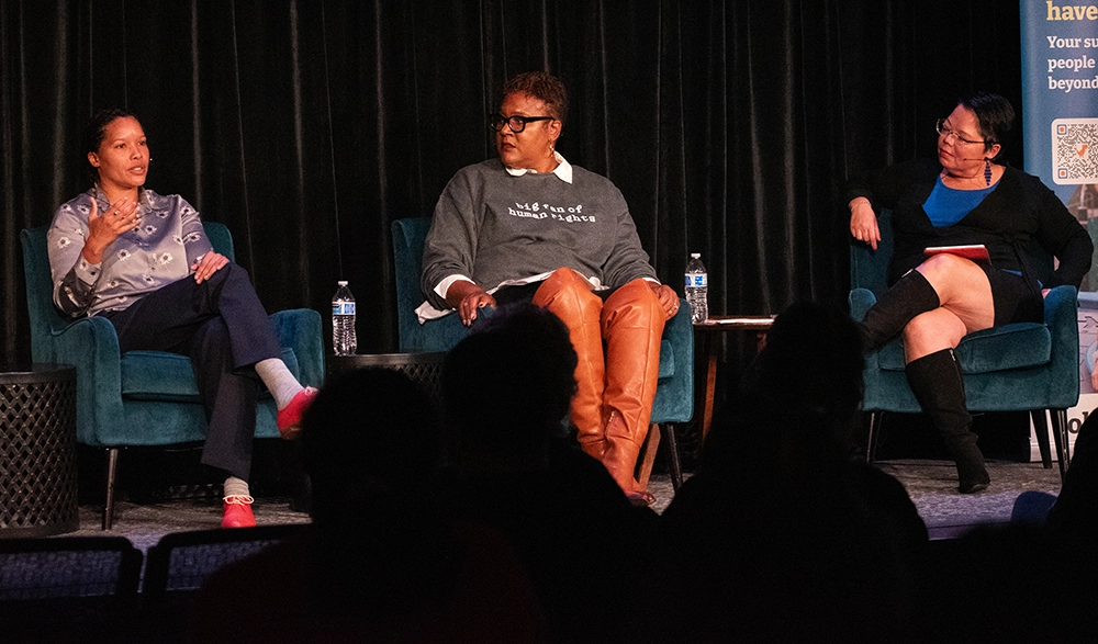 A woman sitting on a dark stage speaks as two others look on.
