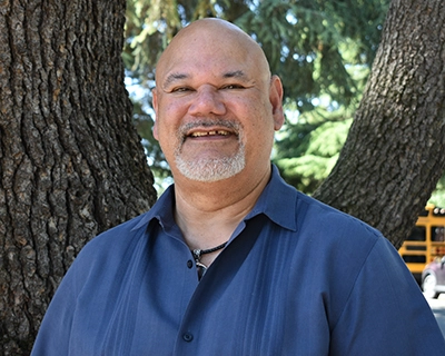 A bald man with a short wh ite beard and a blue shirt smiles as he stands in front of the trunk of a tree